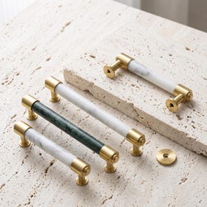 May include: Four cylindrical cabinet pulls featuring polished gold-tone metal end caps and mounts. Three pulls have white marble bodies with grey veining, and one pull has a dark green marble body. A single circular gold-tone metal washer is included.