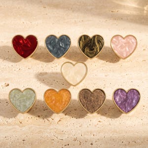 May include: A collection of nine heart-shaped knobs with gold-toned metal rims. The centres feature varied marbled patterns in red, dark blue, metallic gold, light pink, cream, pale green, orange, brown, and purple, arranged in three rows.