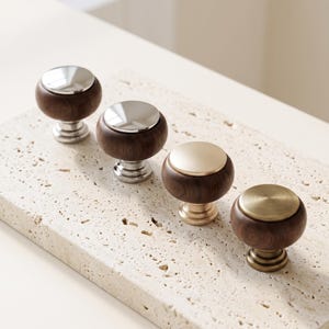 Op de afbeelding: Vier kastknoppen met een afgerond donkerbruin houten lichaam en een ronde metalen bovenkant. Twee knoppen hebben gepolijste zilverkleurige metalen bovenkanten en voetjes, terwijl de andere twee geborstelde goudkleurige metalen afwerkingen hebben.