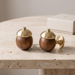 May include: Two spherical wall hooks featuring dark brown wood bases and brushed brass caps. One hook is shown from the front, while the other is attached to a circular brass mounting plate, displaying the hardware used for wall installation.