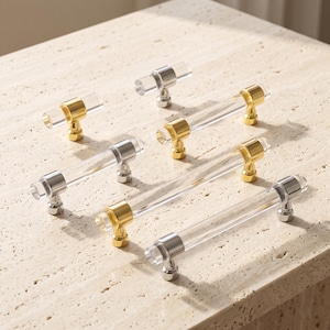 May include: A collection of six cabinet pulls and knobs featuring clear cylindrical acrylic bars accented with polished gold-tone metal mounting posts. The hardware set includes various lengths, each with a transparent body and metallic ends.