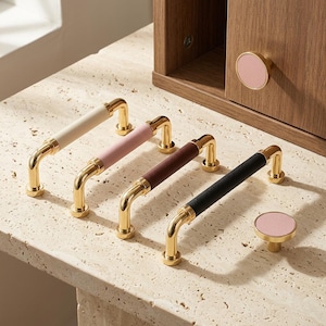 Puede incluir: Cuatro tiradores de cajón de metal dorado con agarres centrales en crema, rosa claro, marrón oscuro y negro, junto con dos pomos redondos dorados con inserciones circulares rosas, uno de los cuales está fijado a un panel de puerta de madera.