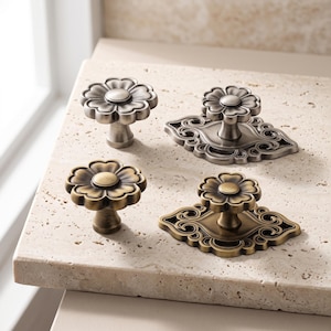 May include: Four metal cabinet knobs in two finishes, silver and antique brass. Two knobs are simple flower shapes, while the other two feature the same flower design mounted on an ornate, elongated backplate with intricate scrollwork details.