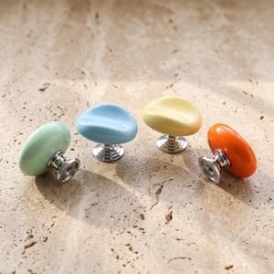 Op de afbeelding: Vier kastknoppen met gladde, afgeronde, kiezelvormige bovenkanten in pastelgroen, lichtblauw, lichtgeel en feloranje. Elke knop is voorzien van een gepolijste zilverkleurige metalen voet met een ronde montageplaat en een schroefgat.