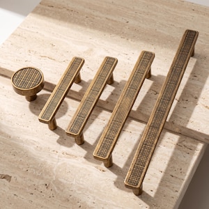 May include: A collection of five antique brass cabinet hardware pieces, including one round knob and four linear pulls of varying lengths. Each piece features a recessed, black grid pattern inlay along the center of the metallic surface.