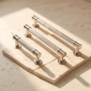 May include: Three silver-toned metal cabinet pulls with a cylindrical, vertically ribbed design. Each handle features two circular mounting bases at the ends, showcasing a polished, reflective finish that catches the light across the ridges.