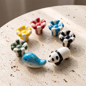 May include: A collection of ceramic objects including five flower-shaped knobs with striped patterns in red, blue, yellow, green, and black, alongside a blue whale figurine and a black and white panda figurine arranged in a circular formation.
