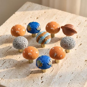 Puede incluir: Una colección de diez pomos decorativos para armarios con forma de planetas y lunas, cada uno con una superficie texturizada única en colores azul, naranja, gris y marrón, montados sobre pequeñas bases cilíndricas de metal dorado.