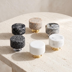 Puede incluir: Una colección de seis pomos circulares para armarios con diversas texturas de piedra, incluyendo patrones de granito negro, blanco y moteado. Cada pomo está montado sobre una base metálica, con acabados en tonos dorados y plateados.