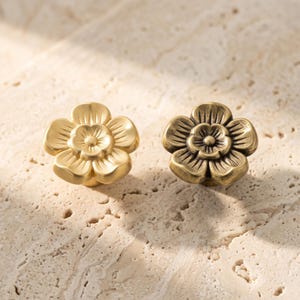 May include: Two flower-shaped cabinet knobs with five rounded petals and a central circular detail. One knob features a smooth, matte gold finish, while the other has an antiqued, brushed bronze finish with darker recessed areas between the petals.