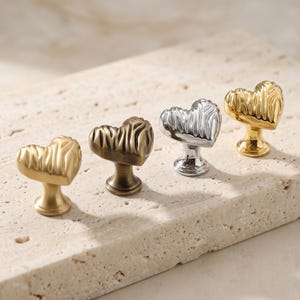 May include: Four heart-shaped cabinet knobs with a textured, rippled surface. The set includes one matte gold knob, one antique bronze knob, one polished silver knob, and one polished gold knob, each featuring a circular base for mounting.