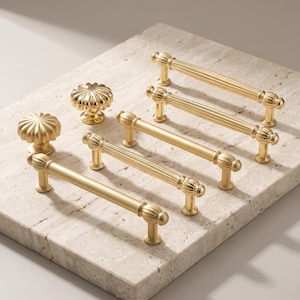 May include: A collection of gold-toned metal cabinet hardware, including two round fluted knobs and four drawer pulls. The pulls feature a mix of smooth and vertically ribbed cylindrical bars with decorative fluted end caps and mounting posts.