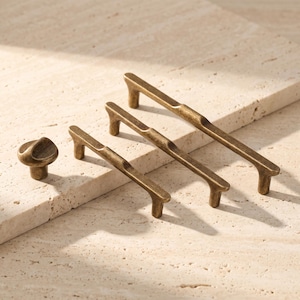 May include: A set of three antique brass cabinet hardware pieces consisting of one round knob and two elongated drawer pulls. Each piece features a smooth, curved design with a metallic finish, showcasing a consistent aged bronze colour tone.