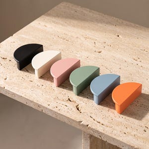 Könnte beinhalten: Ein Set aus sechs halbkreisförmigen Schrankgriffen, die in einer Reihe angeordnet sind. Die Griffe haben eine matte Oberfläche in den Farben Schwarz, Creme, Altrosa, Salbeigrün, Schieferblau und leuchtendem Orange mit flacher Vorderseite.