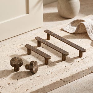 May include: A collection of antique brass cabinet hardware including two round knobs with a radial sunburst pattern and three rectangular bar pulls featuring a textured, linear grooved surface and cylindrical mounting posts for attachment.