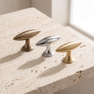 Peut inclure: Trois boutons de placard en forme de T avec des extrémités allongées et pointues. Un bouton présente une finition en laiton antique brossé, le deuxième une finition chromée polie et le troisième une finition dorée mate et lisse.