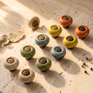 Peut inclure: Un ensemble de onze boutons de placard ronds en céramique de différentes couleurs, notamment orange, jaune, bleu et vert. Chaque bouton présente une finition émaillée mouchetée et une incrustation centrale en métal bronze en forme de fleur.