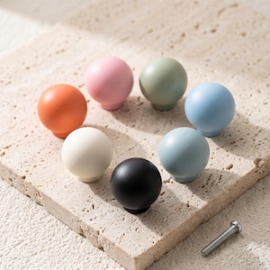 May include: A collection of seven spherical cabinet knobs in various solid matte colors including orange, pink, sage green, light blue, cream, black, and dusty blue, arranged in a circular pattern with a single metal mounting screw nearby.