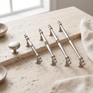 May include: A set of brushed nickel cabinet hardware including one round knob and four elongated drawer pulls. Each piece features a smooth, metallic finish with a tapered design and rounded ends, providing a consistent look for furniture.