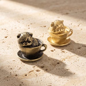 Op de afbeelding: Twee miniatuur metalen beeldjes, elk met een kat die in een theekopje op een schoteltje zit. Het ene beeldje heeft een donkere, antieke bronskleurige afwerking, terwijl het andere een heldere, gepolijste gouden afwerking heeft.