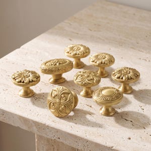 May include: A collection of eight brass-toned cabinet knobs featuring various embossed designs, including floral patterns, an oval shape with leaf motifs, and a circular knob with a geometric Greek key border, all with a matte metallic finish.