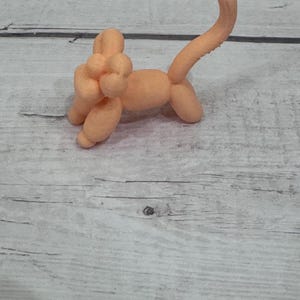 May include: A peach-colored balloon animal in the shape of a dog. The balloon dog has a long tail, four legs, and a head with a small knot. The dog is standing on a light gray wooden surface.