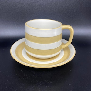 Puede incluir: Un juego de taza y platillo de café de cerámica. La taza y el platillo están decorados con rayas horizontales alternas de color blanco y amarillo-marrón apagado. La taza tiene un asa a juego.