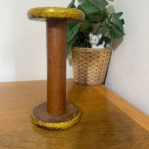 Può includere: Un rocchetto di legno vintage con un bordo giallo sopra e sotto. Il rocchetto ha un centro cilindrico marrone e poggia su una superficie di legno. Una piccola statuina di ceramica e una pianta in vaso sono sullo sfondo.