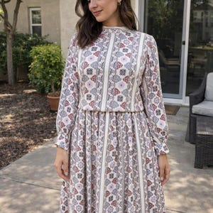 May include: A long-sleeved, floor-length dress with a white background and a repeating pattern of vertical stripes and floral motifs in red, purple, and grey. The dress has a high neckline and a fitted waist. The model is standing outside.