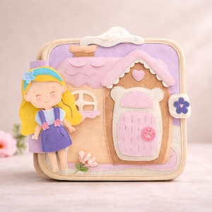May include: A felt dollhouse with a smiling doll figure. The dollhouse is beige, pink, and purple with a chimney, a heart, and a flower-shaped button. The doll has yellow hair, a blue headband, and a purple dress.