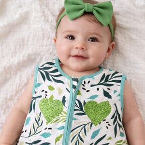 May include: A baby in a sleeveless sleep sack featuring a botanical print in green and blue hues. The sleep sack has a teal zip and two green glitter heart appliqués. The baby is also wearing a green bow headband.