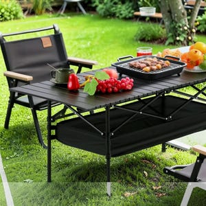 Puede incluir: Una mesa de camping plegable negra con una silla negra y una parrilla encima. La mesa está puesta con comida y bebida, incluyendo uvas, un pimiento rojo y una taza de café. La mesa está colocada sobre un césped verde.