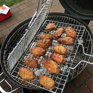 Könnte beinhalten: Ein schwarzer, runder Holzkohlegrill mit einem Metallrost, auf dem mehrere gegrillte Hähnchenflügel liegen. Die Flügel sind goldbraun und sehen frisch gegrillt aus. Der Grill steht im Freien.