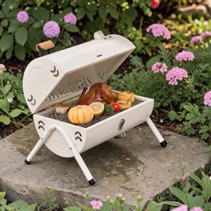 May include: A white, retro-style grill with the lid open, revealing cooked food. The grill has a wooden handle and is set on a stone surface surrounded by flowers. The grill contains a roasted chicken, vegetables, and a small pumpkin.