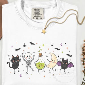 May include: A white t-shirt featuring a graphic print of a black cat, a skeleton, a potion bottle with green liquid, a crescent moon, and a bat with purple wings. The shirt has a Comfort Colors label at the neck and a gold star pendant necklace.