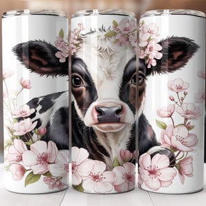 May include: A white tumbler featuring a black and white cow with a floral crown and pink flowers. The cow's face is centered on the tumbler, surrounded by delicate blossoms. The design is repeated on all three tumblers.