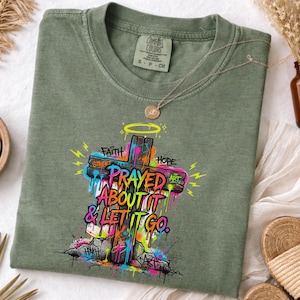 May include: Sage green t-shirt featuring a colorful graffiti-style cross with the words "Prayed About It & Let It Go." The cross is adorned with the words "Faith," "Hope," and "Art." A gold necklace with a pendant rests on the shirt.