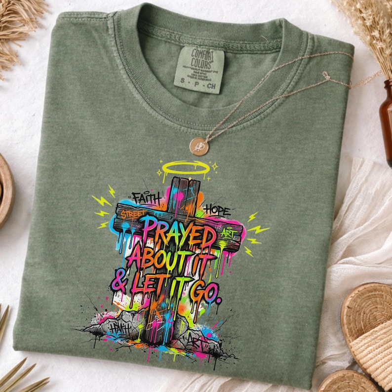 May include: Sage green t-shirt featuring a colorful graffiti-style cross with the words "Prayed About It & Let It Go." The cross is adorned with the words "Faith," "Hope," and "Art." A gold necklace with a pendant rests on the shirt.