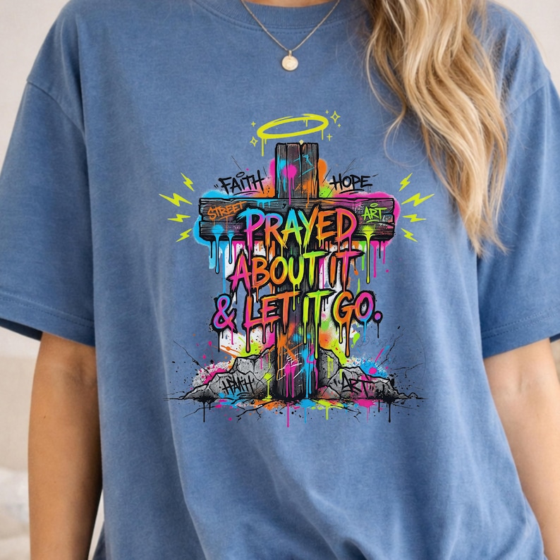 May include: Blue t-shirt featuring a colorful graphic of a wooden cross with the words "Prayed About It & Let It Go." The design includes the words "Faith," "Hope," "Street," and "Art" with a halo and lightning bolts.