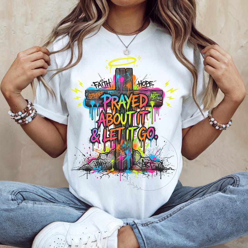 May include: White t-shirt featuring a colorful graffiti-style cross design. The cross has the words "Faith" and "Hope" on either side, with "Prayed About It & Let It Go" in the center. The design includes dripping paint and a halo.
