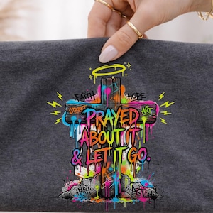 May include: A dark gray t-shirt featuring a colorful graffiti-style cross design. The cross has the words "Prayed About It & Let It Go" and other words like "Faith" and "Hope". The design includes dripping paint and lightning bolts.
