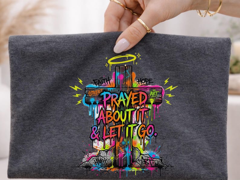 May include: A dark gray t-shirt featuring a colorful graffiti-style cross design. The cross has the words "Prayed About It & Let It Go" and other words like "Faith" and "Hope". The design includes dripping paint and lightning bolts.