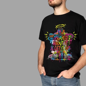 May include: Black t-shirt featuring a colorful graffiti-style cross design. The cross has the words "PRAYED ABOUT IT & LET IT GO" in a vibrant, multi-colored font. A yellow halo and lightning bolts are also included in the design.