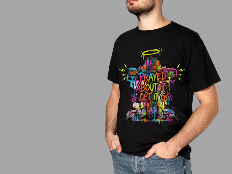 May include: Black t-shirt featuring a colorful graffiti-style cross design. The cross has the words "PRAYED ABOUT IT & LET IT GO" in a vibrant, multi-colored font. A yellow halo and lightning bolts are also included in the design.
