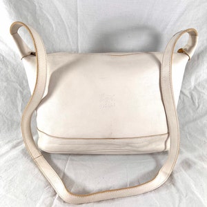 May include: A white leather shoulder bag with a long strap. The bag has a zipper closure and a small logo on the front that says "Country" in a cursive font.