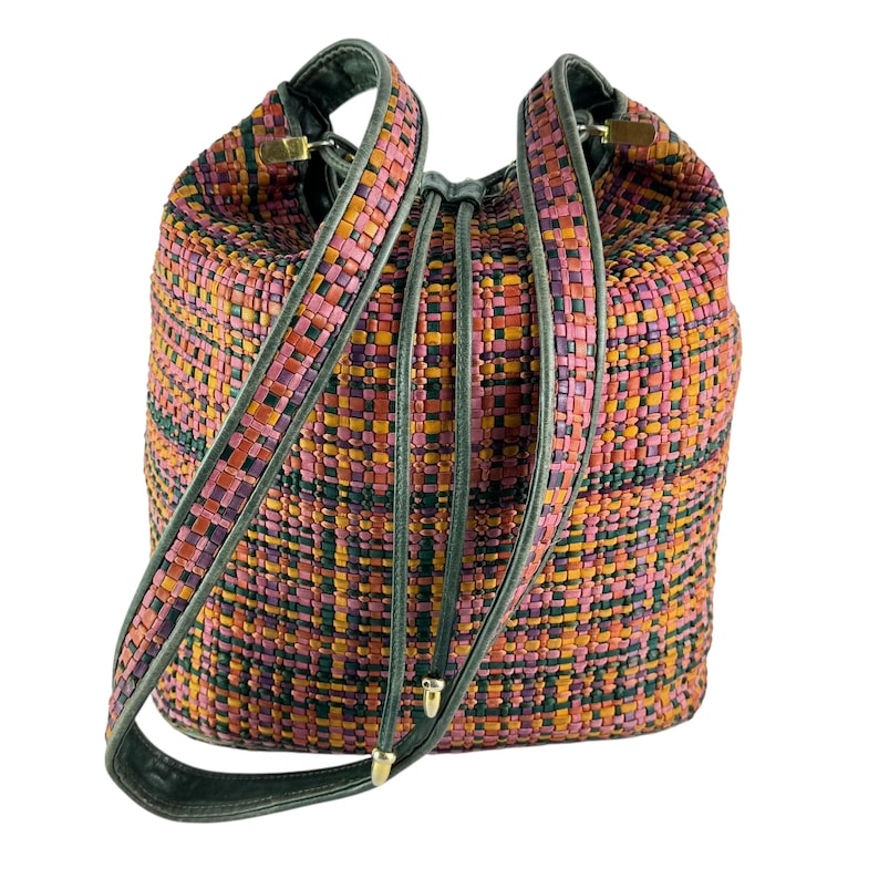 May include: A colourful woven bucket bag with pink, orange, and green hues. It has a drawstring closure and long shoulder straps with dark green trim. Gold-toned hardware accents the straps.