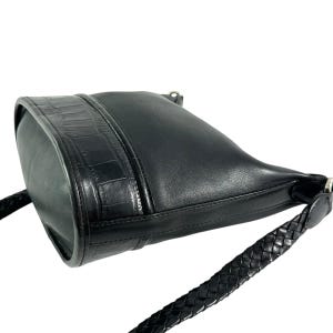 May include: Black leather crossbody bag with a braided strap. The bag features a textured crocodile-style panel on the front and a smooth leather body. The strap is a thick, braided design. The bag is angled, showing its shape and details.