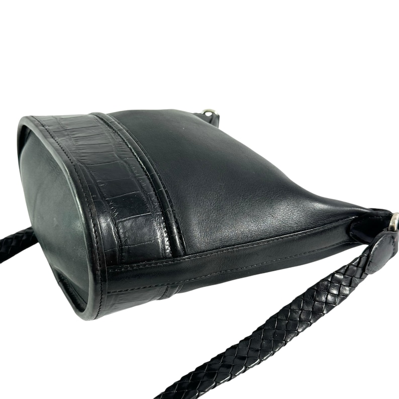 May include: Black leather crossbody bag with a braided strap. The bag features a textured crocodile-style panel on the front and a smooth leather body. The strap is a thick, braided design. The bag is angled, showing its shape and details.