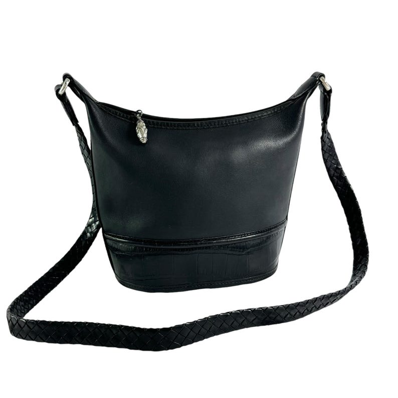 May include: Black leather bucket bag with a braided shoulder strap. The bag features a textured bottom section and a silver-tone charm. The strap is adjustable. The bag is designed to be worn over the shoulder.