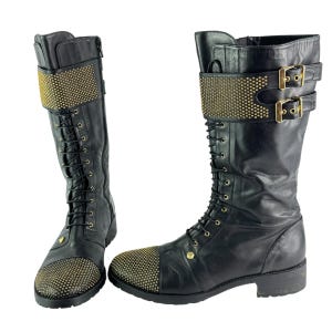 May include: Pair of black leather knee-high boots with gold-colored stud accents. The boots feature a lace-up design, a studded toe cap, and a studded strap with two buckles. The boots have a zipper on the side.